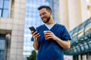 Young male nurse using phone outside while drinking coffee